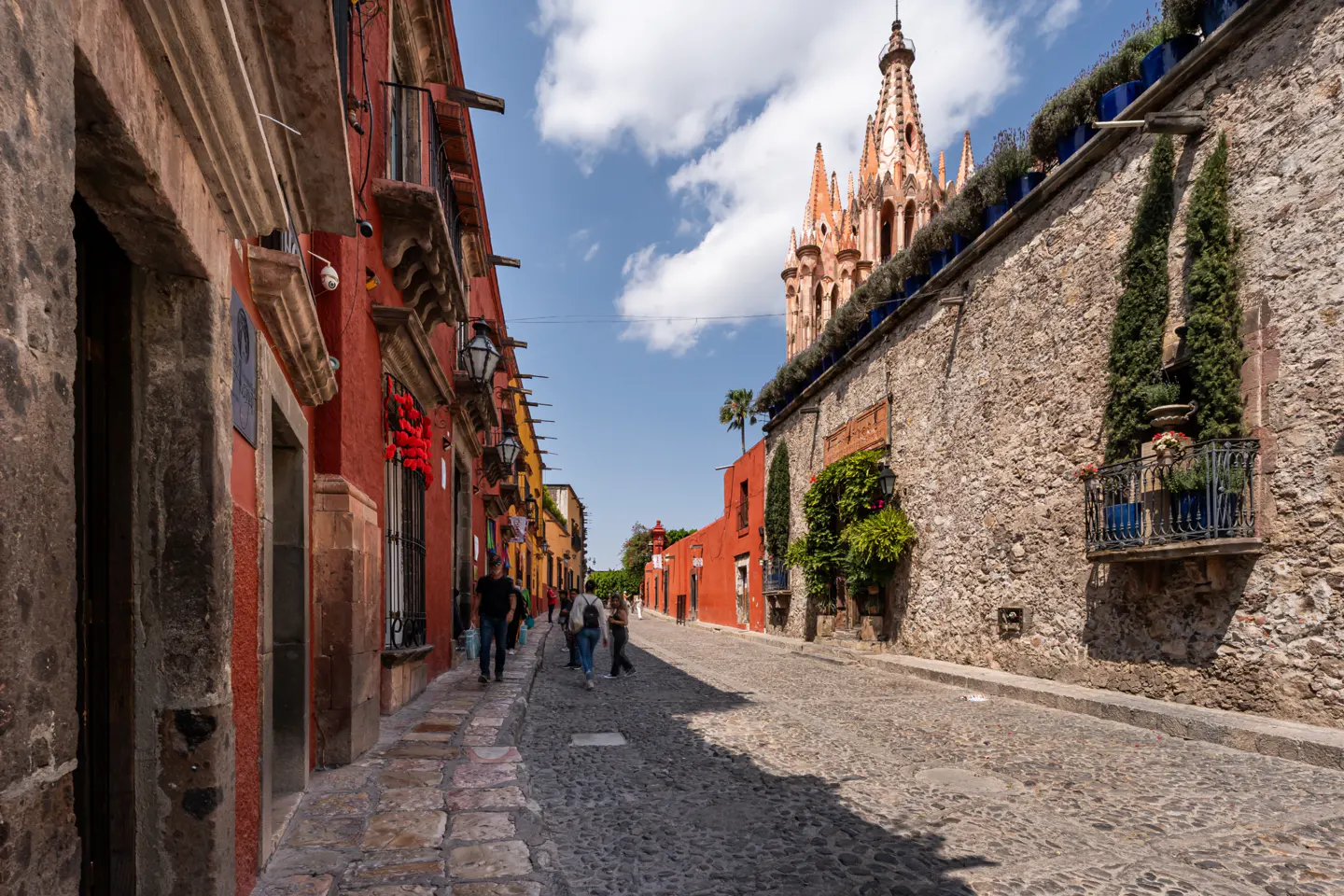 Cobblestone street view in San Miguel de Allende, Mexico. Red buildings line the left, stone wall on the right, church in the background.