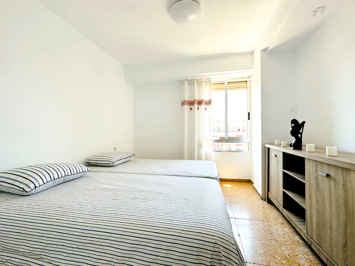 Bright bedroom with two twin beds, striped bedding, and a light wood cabinet with decor near a window.
