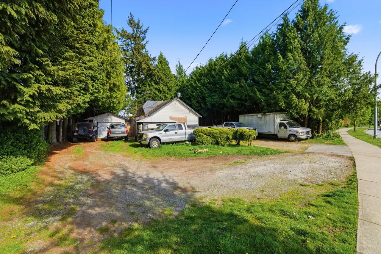 A gravel lot with parked vehicles, including a truck and van, surrounded by green trees and grass under a blue sky.