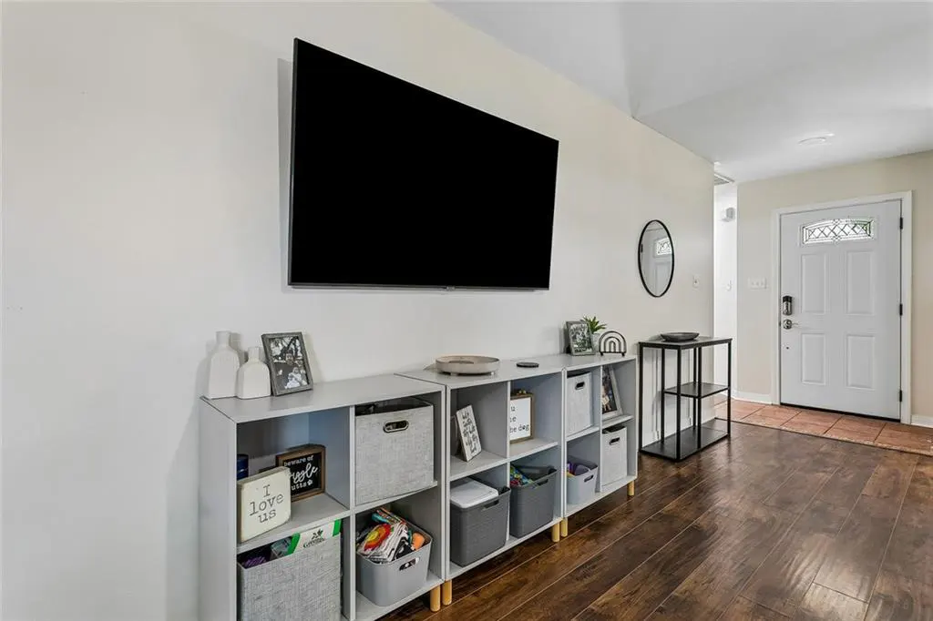 Living room with a large TV mounted above a white shelving unit. Dark wood floors and a white front door are visible.