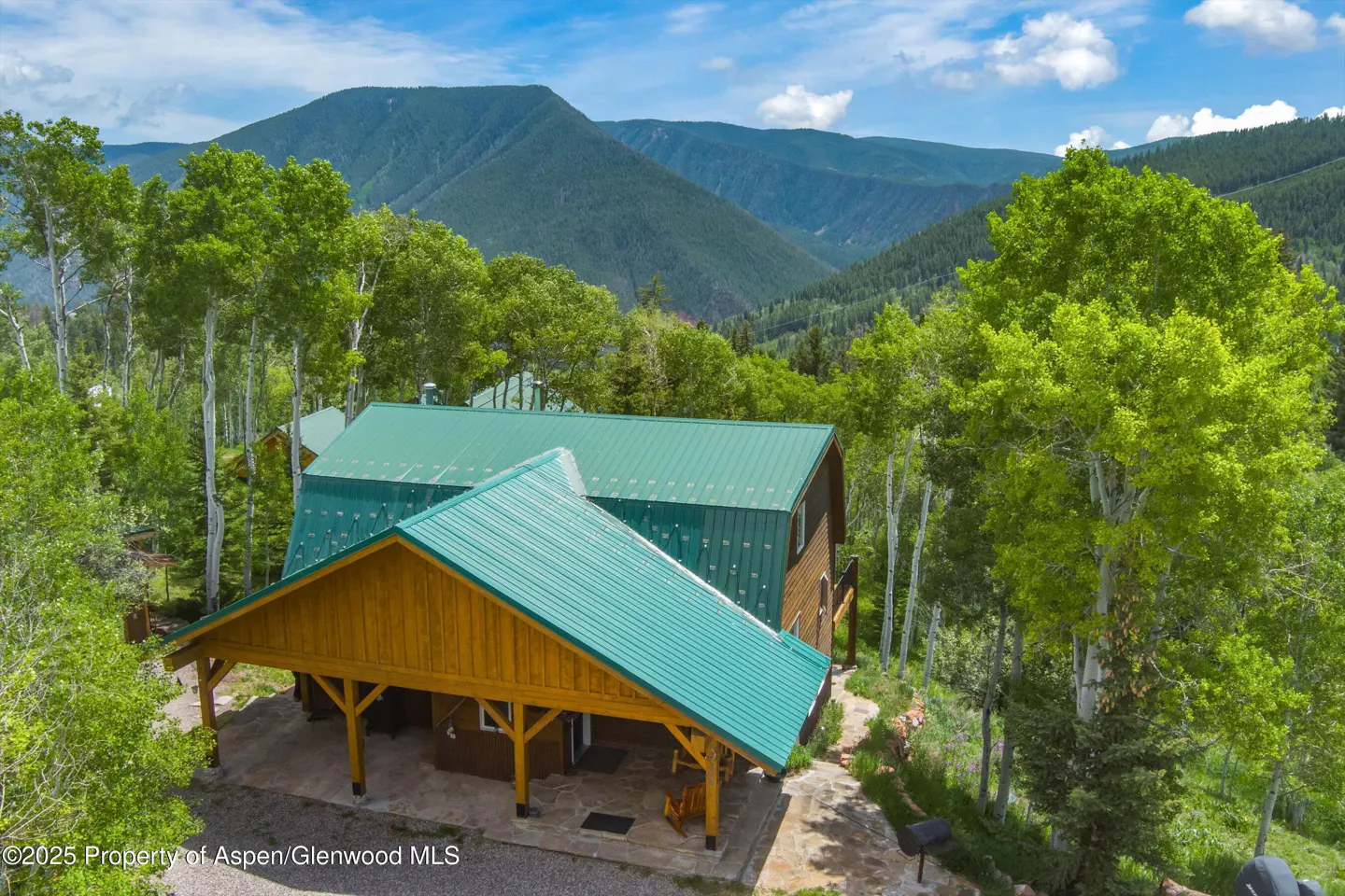 Cabin with a green metal roof and wood beams, surrounded by green trees and mountains under a blue sky.