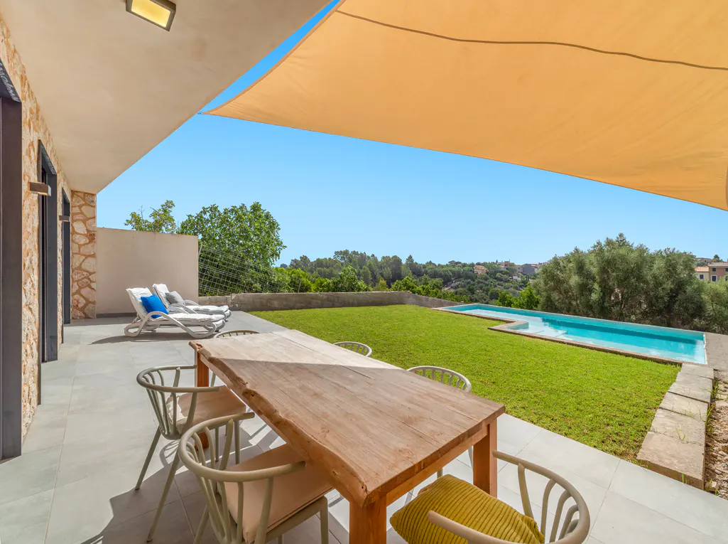 Outdoor patio with a wooden table, chairs, and lounge chairs overlooking a green lawn and pool under a beige sunshade.