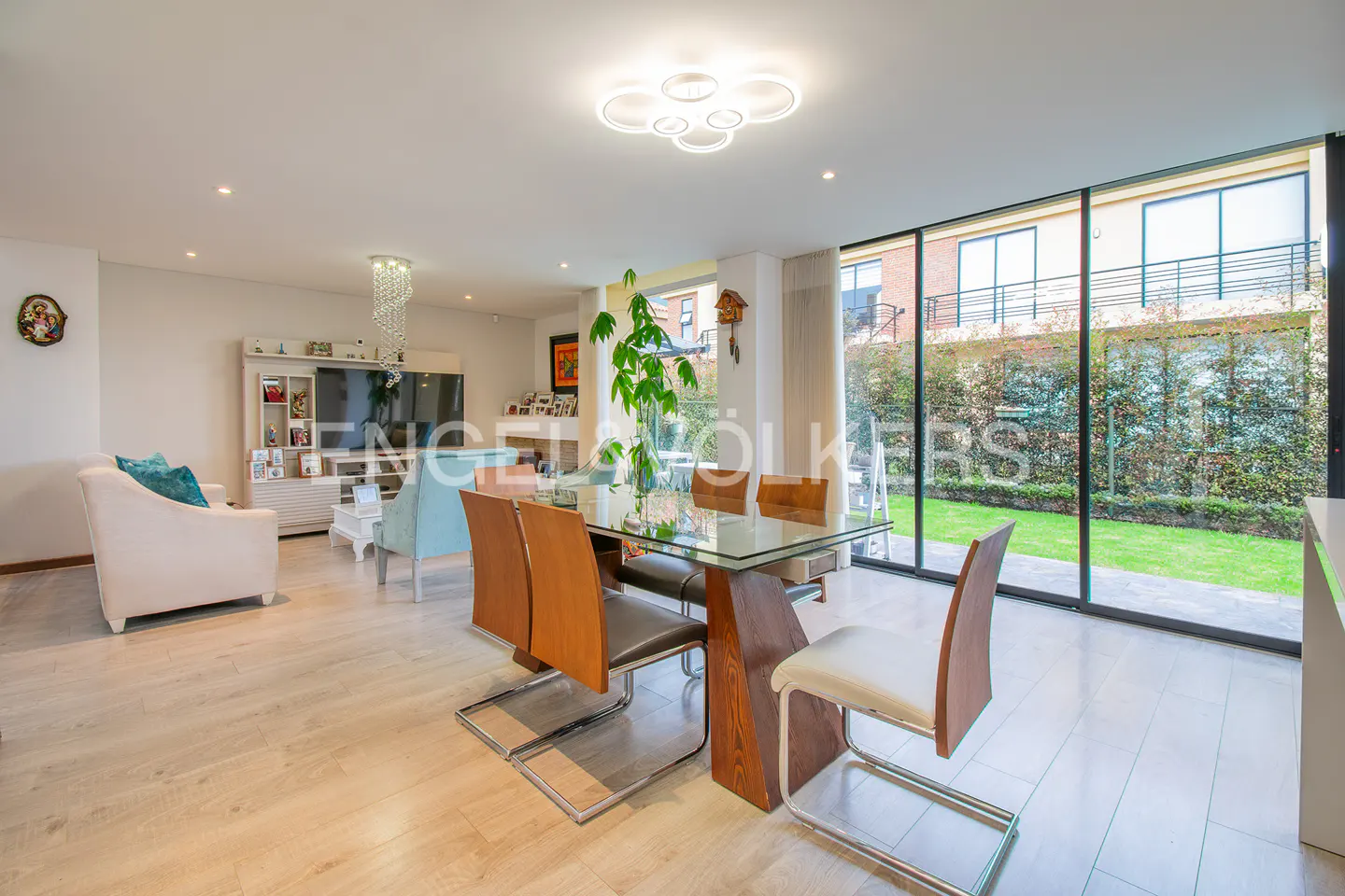 Bright, open-plan living and dining area with a glass table, wood chairs, and a large window overlooking a green garden.