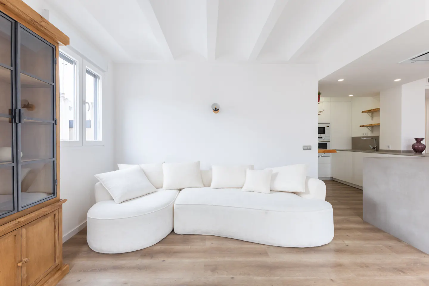 Modern living room with a curved white sofa, wooden cabinet, and an open-plan kitchen with light wood flooring.