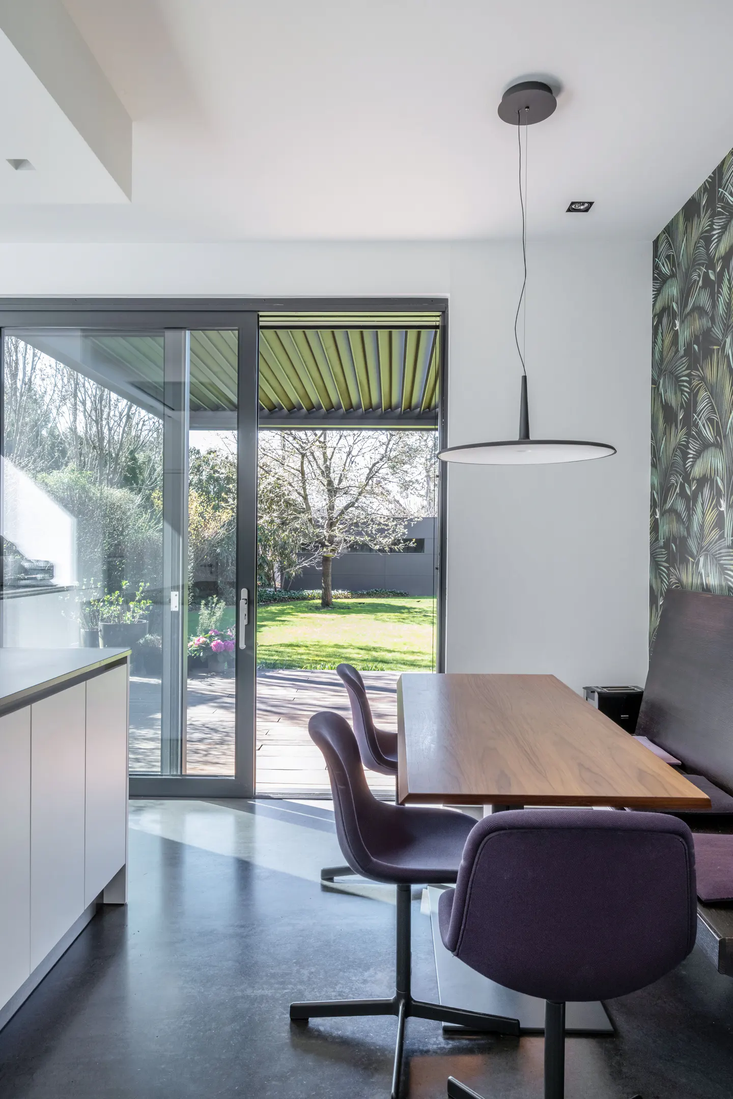 Modern dining area with a wood table, purple chairs, and a bench seat against a palm-leaf wallpapered wall. Sliding glass doors open to a green lawn.