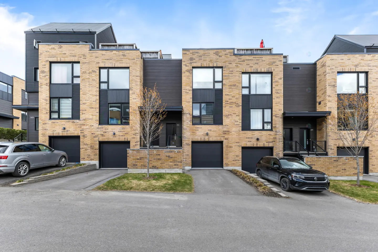 Modern townhouses with tan brick and black siding, each with a garage and large windows, under a blue sky.