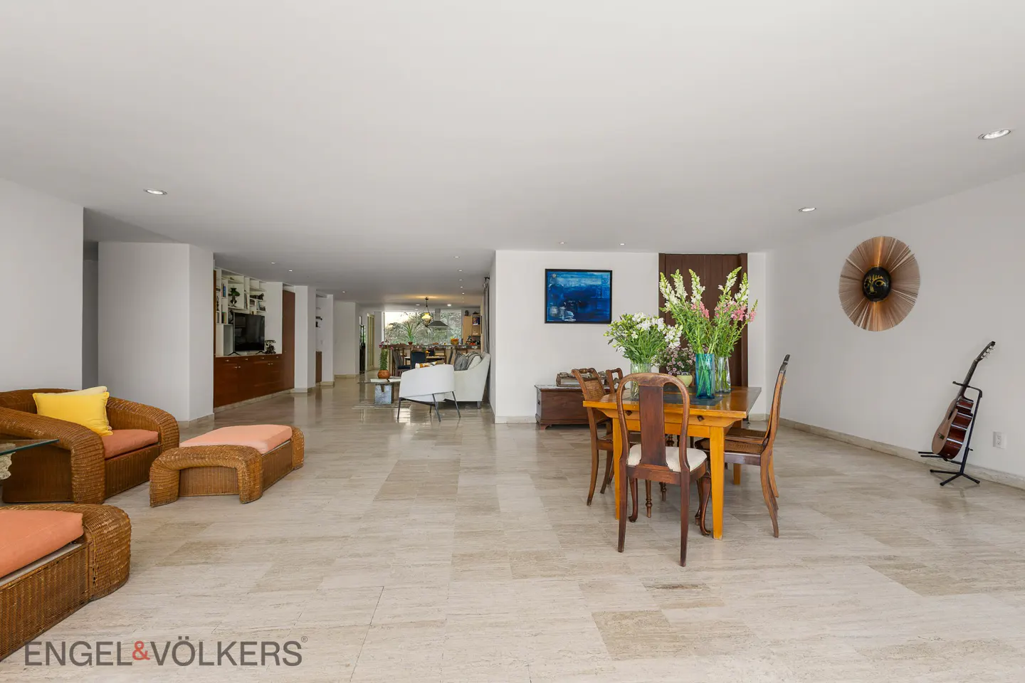 Wide shot of a bright, open-concept living and dining area with wicker furniture, a wooden dining table, and a guitar.