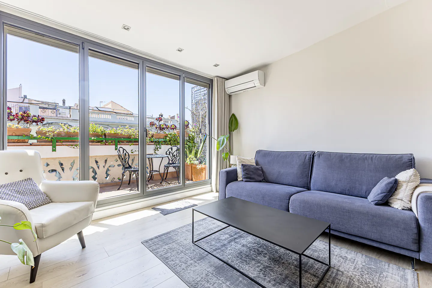 Bright living room with a blue sofa, white armchair, black coffee table, and a balcony with plants and outdoor furniture.