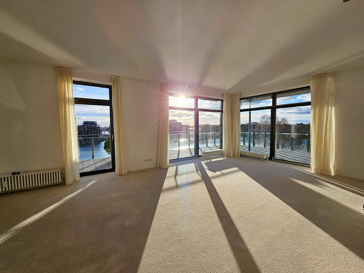 Bright, empty room with beige carpet and white walls. Large windows with cream curtains overlook a river and buildings. Sunlight streams in.