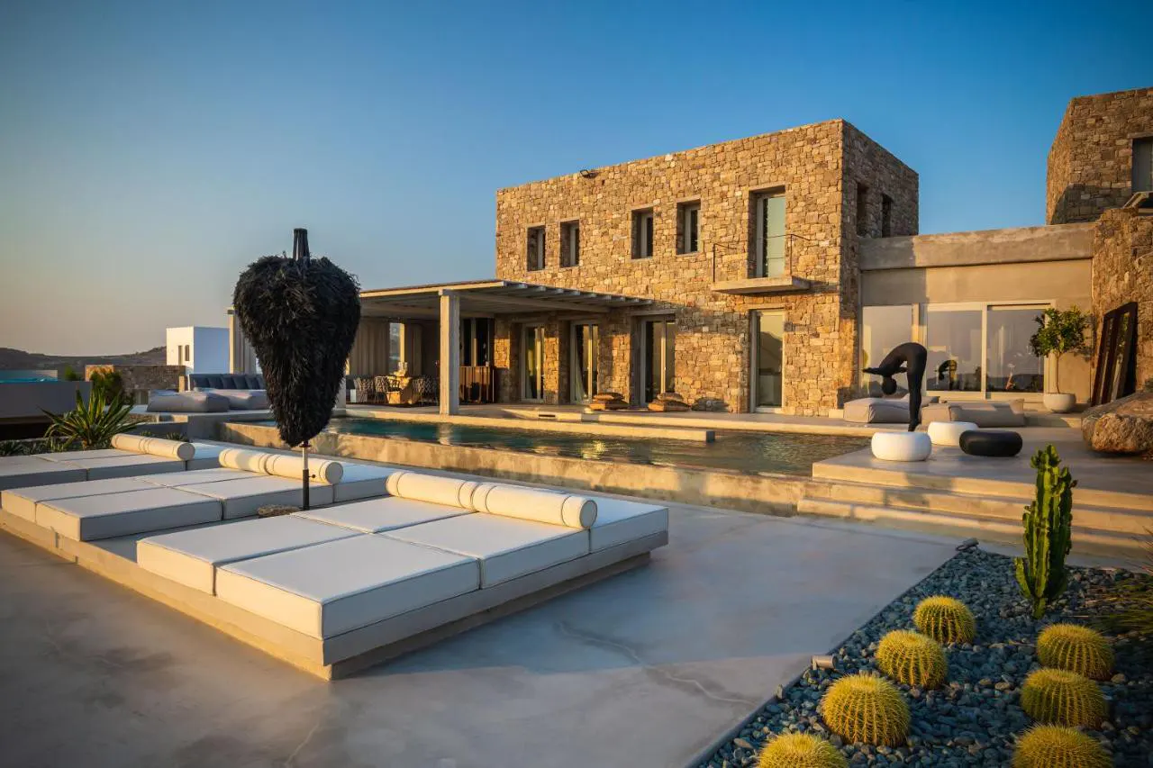 Luxury villa exterior with pool, stone facade, and lounge chairs. A black feather sculpture stands near yellow barrel cacti.