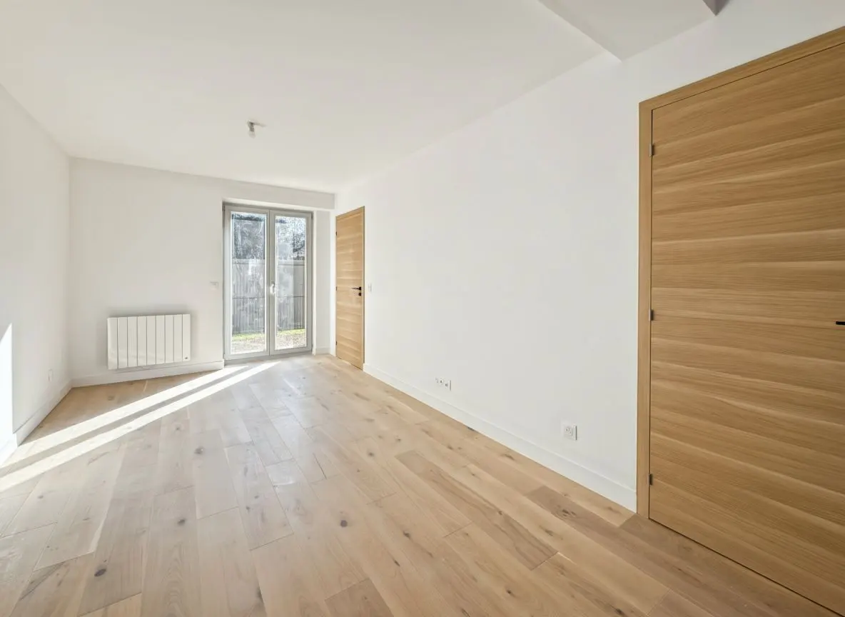 Bright, empty room with light wood floors, white walls, and a wood door. Glass doors lead to a backyard. A white radiator is on the wall.