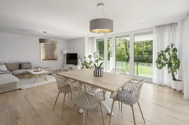 Bright, modern living and dining area with a light wood table, six chairs, and large windows overlooking a green yard.