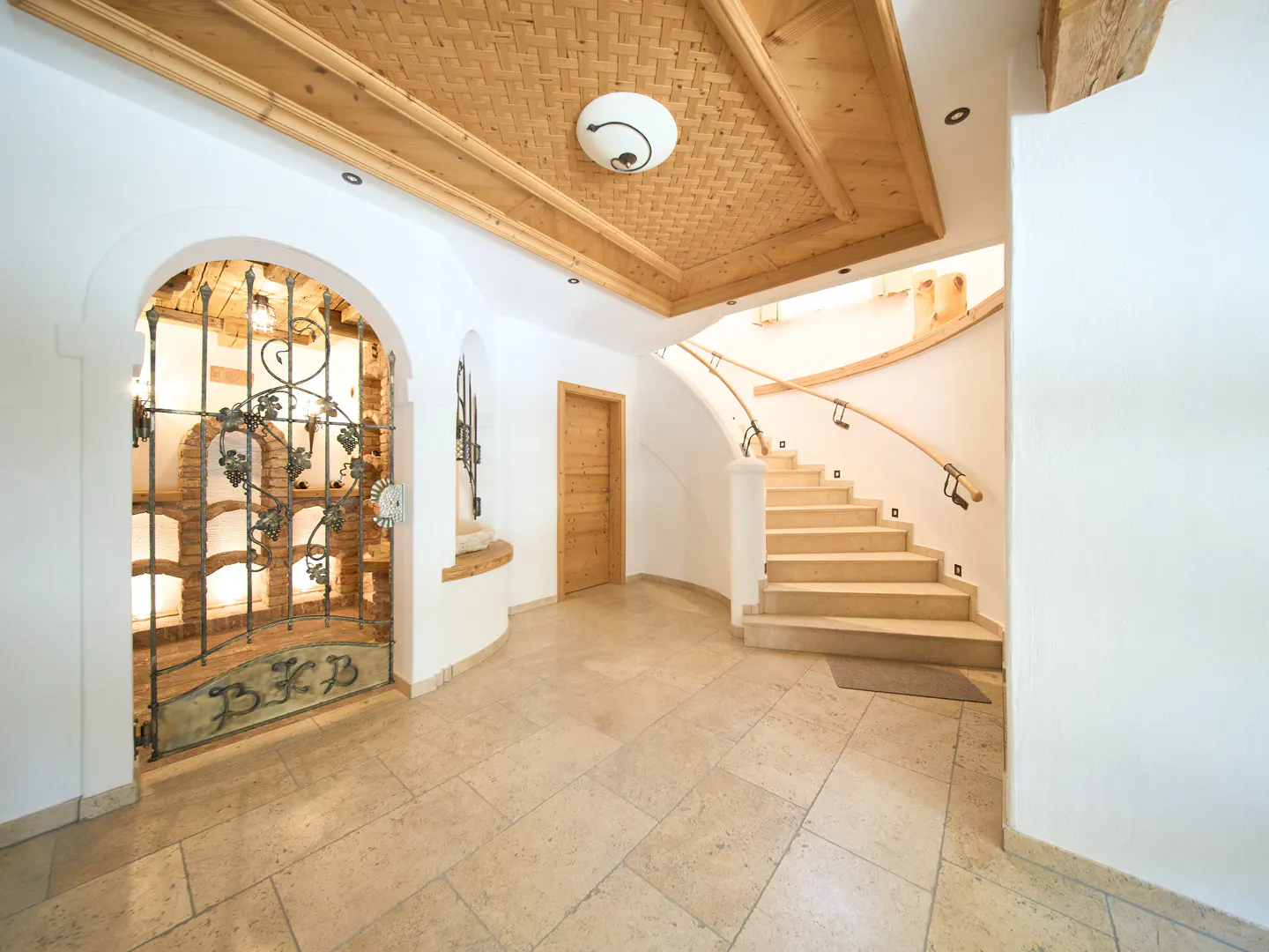 Interior view of a home foyer with stone floors, a wine cellar behind a wrought iron gate, and a curved staircase.