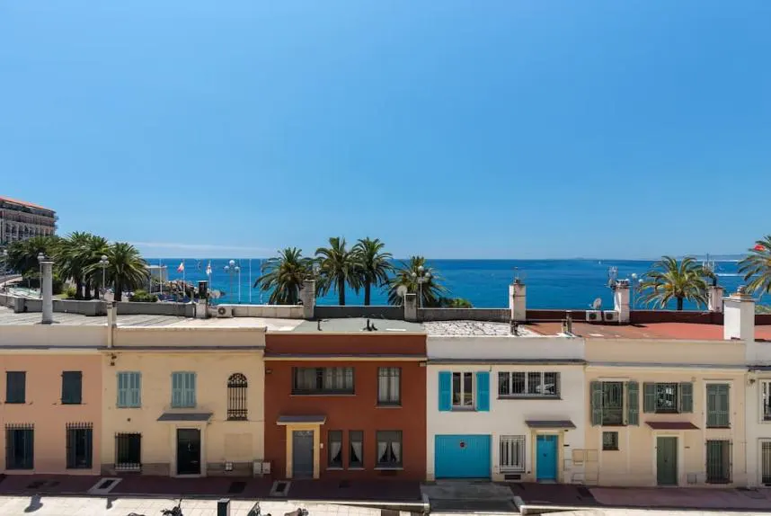 Row of colorful townhouses with blue shutters and doors, overlooking the ocean and palm trees under a clear blue sky.