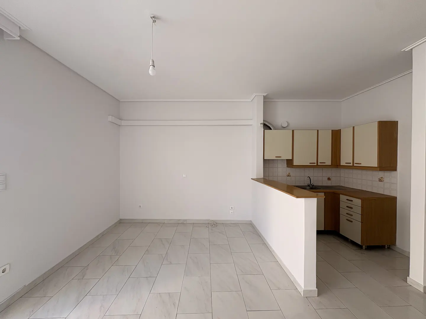 Empty room with white walls, light tile flooring, and a small kitchen area featuring wood-trimmed cabinets and a counter.