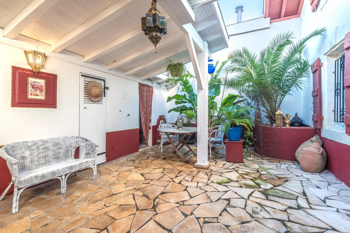 A cozy courtyard with stone flooring, white walls with red trim, plants, and a white wicker bench. Hanging lanterns add a warm glow.