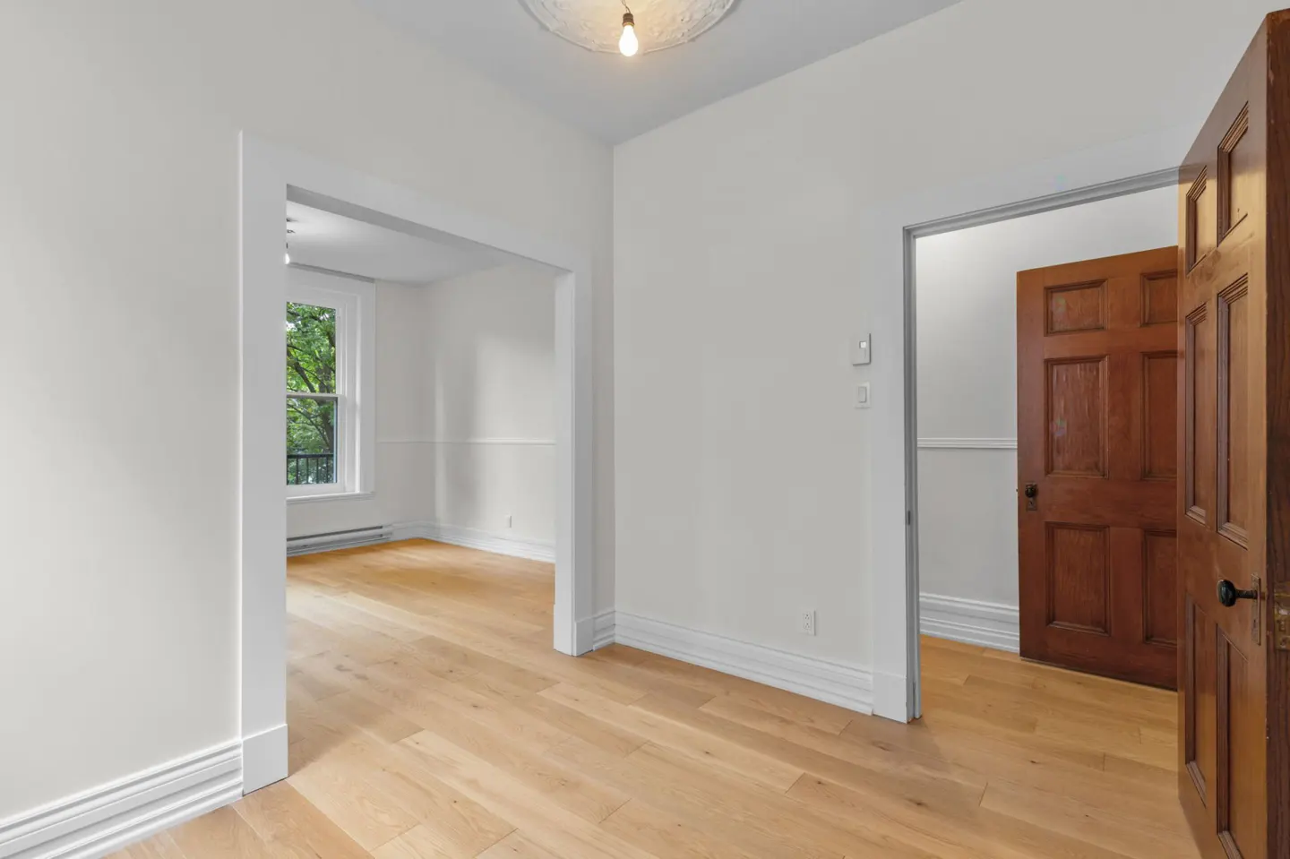 Bright, empty room with light wood floors, white walls, and an open dark wood door. A window is visible in the next room.