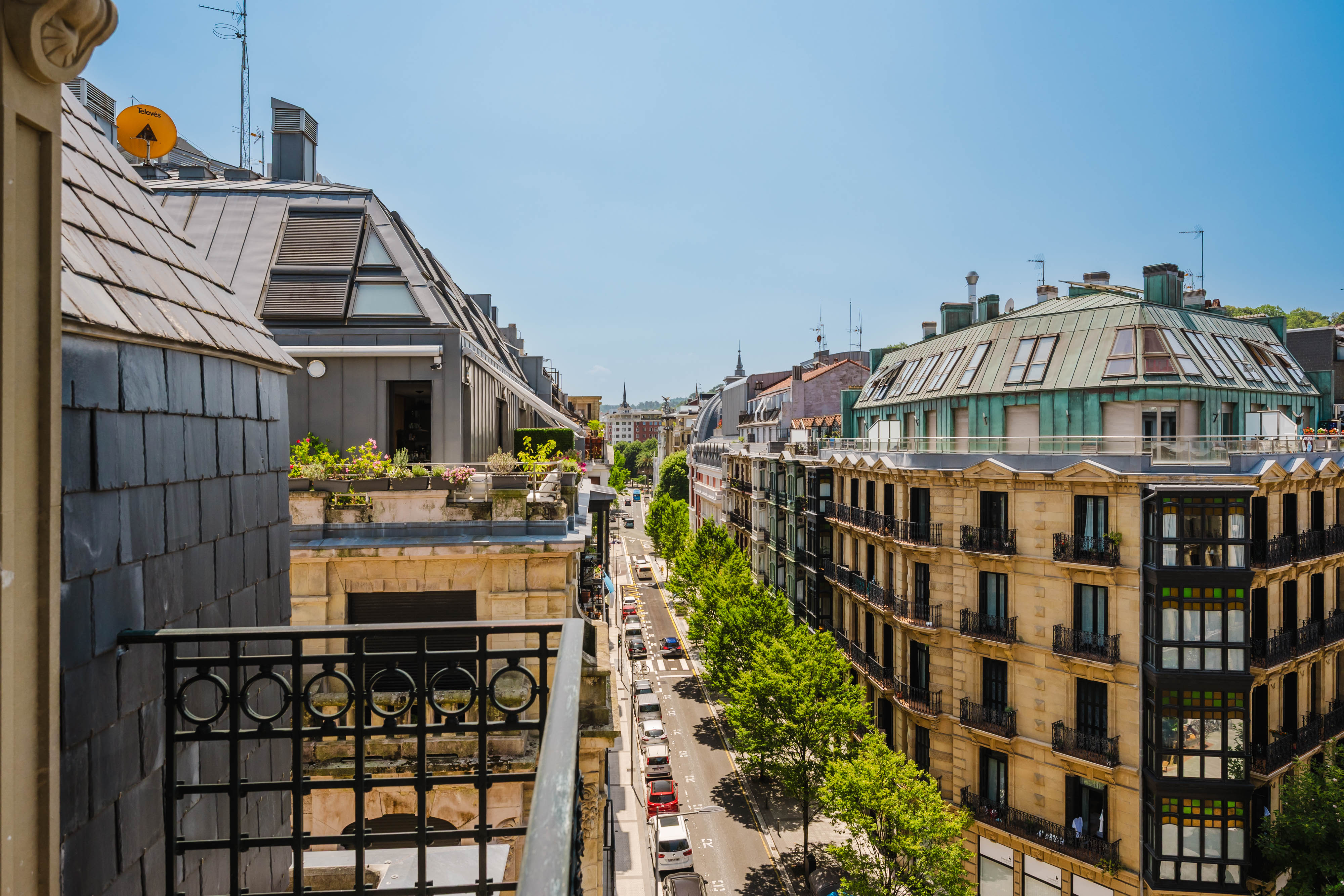 View from a balcony overlooking a sunny European city street lined with trees, historic stone buildings, and parked cars.