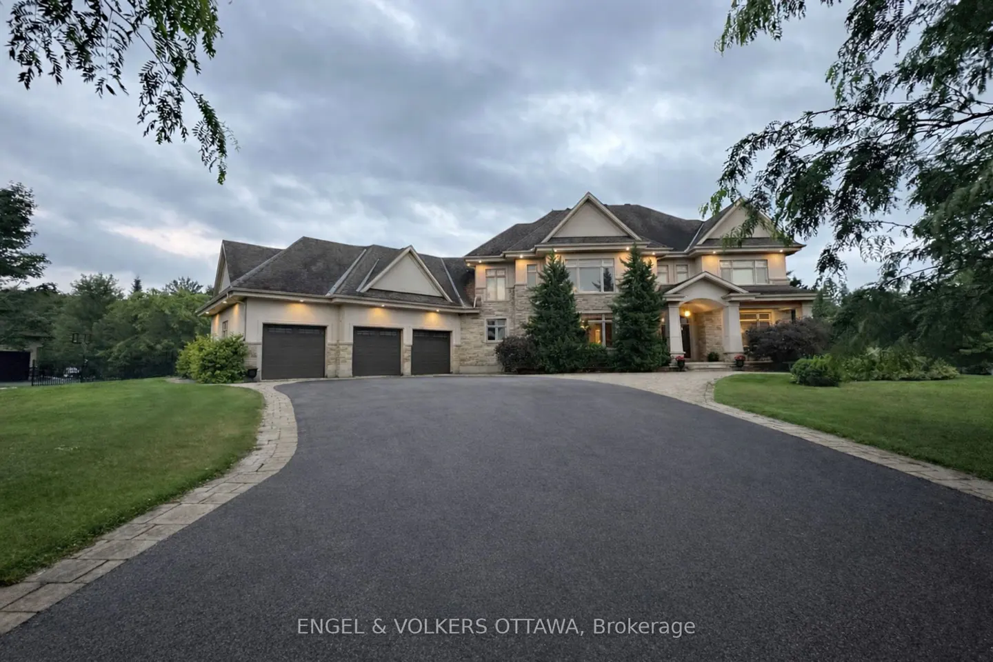 Exterior view of a large, two-story stone house with a three-car garage and a long, paved driveway. Green lawn and trees surround the property.