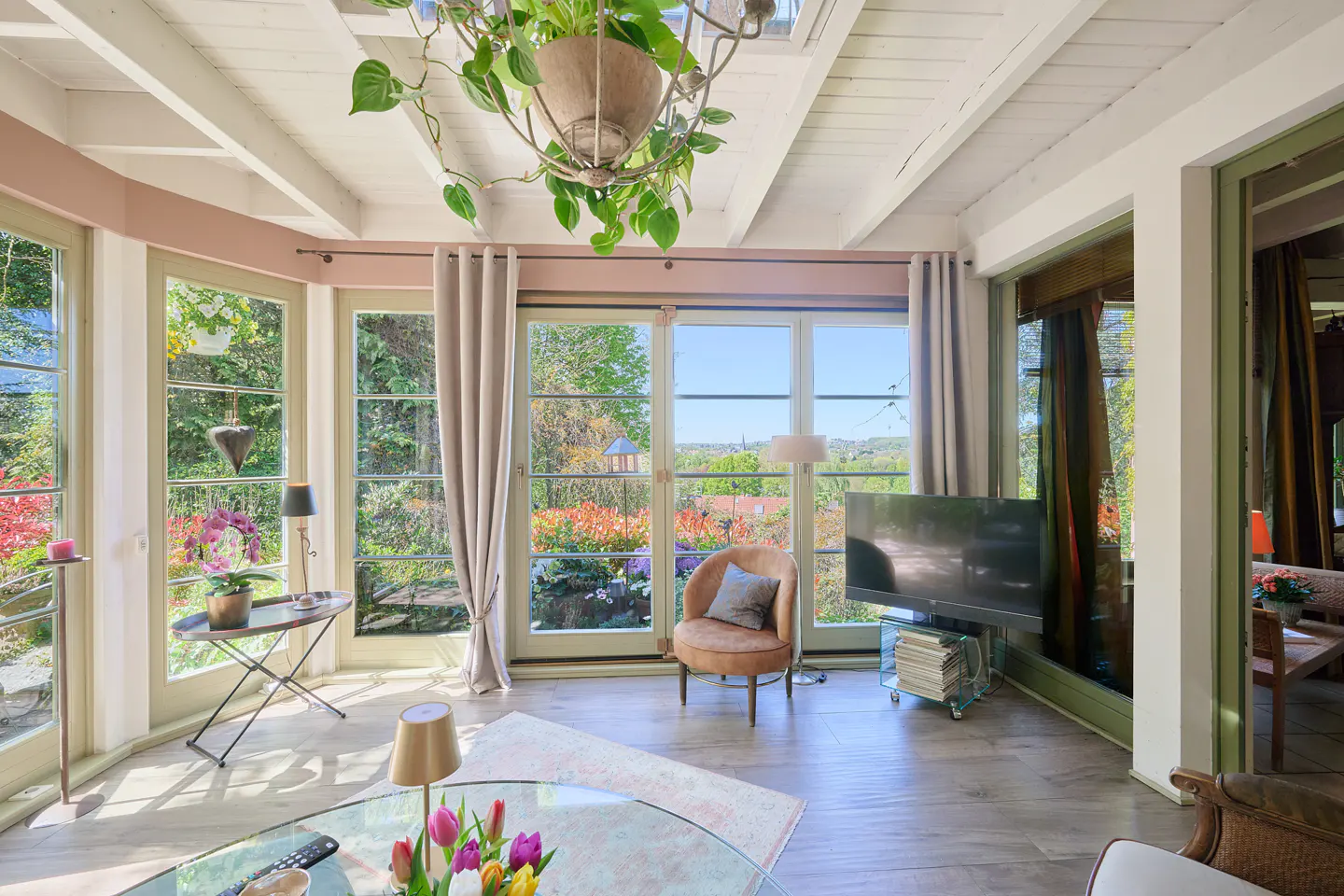 Bright sunroom with white wood ceiling, large windows, and garden views. A chandelier with plants hangs above a glass table with tulips.