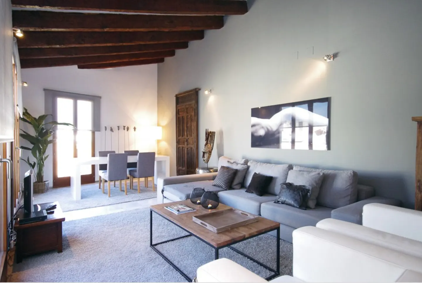 Modern living room with grey sectional sofa, wooden coffee table, exposed ceiling beams, and a dining area in the background.