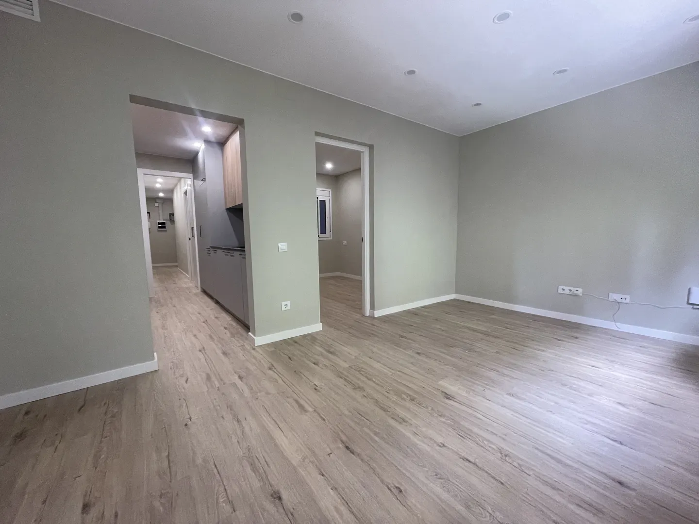 A bright, empty room with light wood floors and gray walls. Doorways lead to a kitchen and another room.