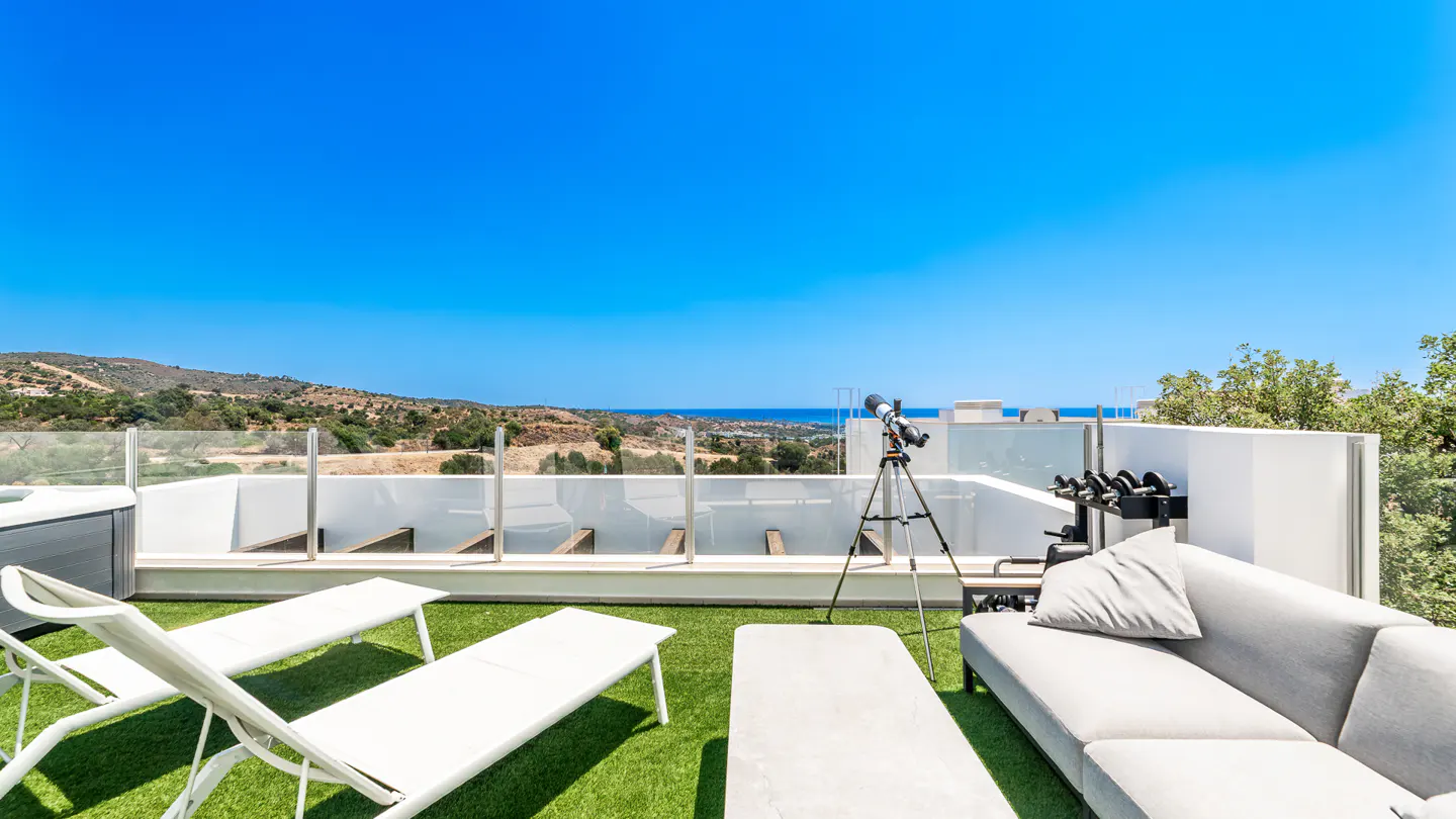 Rooftop patio with white lounge chairs, gray sofa, telescope, and weights on green turf. Blue sky and distant hills in background.
