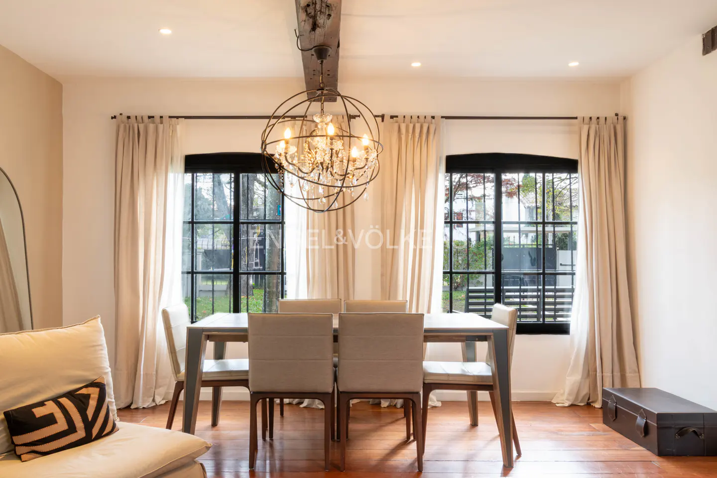 Dining room with a wooden table, six beige chairs, a large orb chandelier, and windows with black frames and curtains.