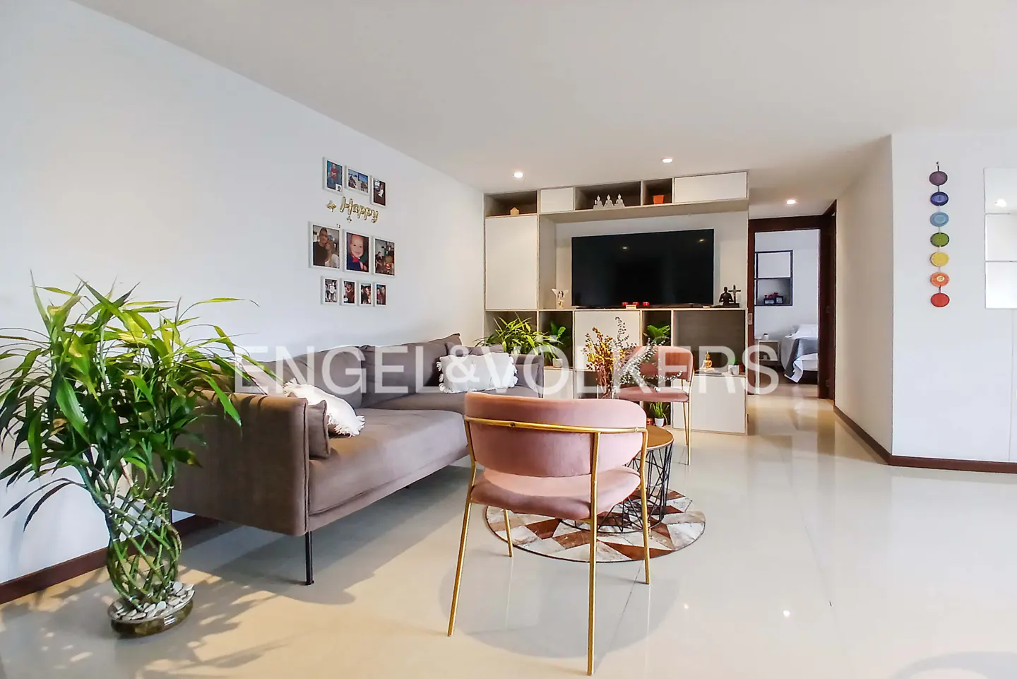 Modern living room with a grey sofa, pink velvet chair, large TV on a shelving unit, and a potted plant on white floors.