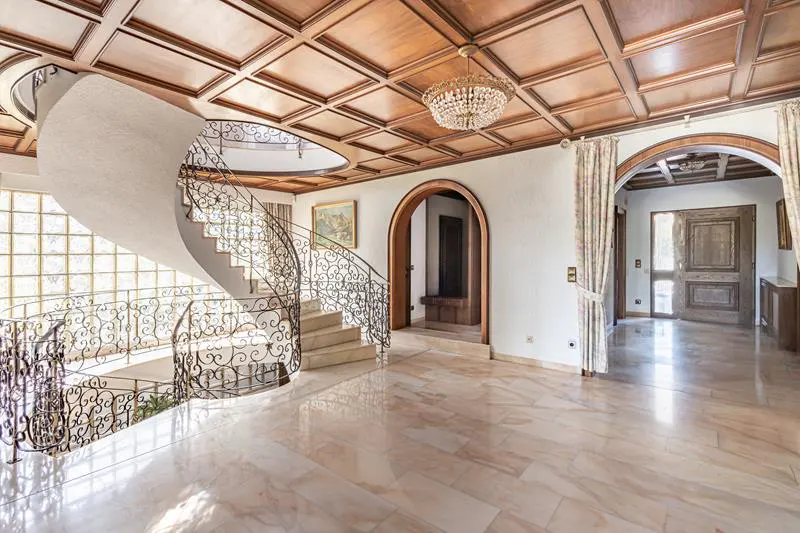 Grand foyer with a spiral staircase, marble floors, and a coffered wood ceiling with a crystal chandelier. Arched doorways lead to other rooms.