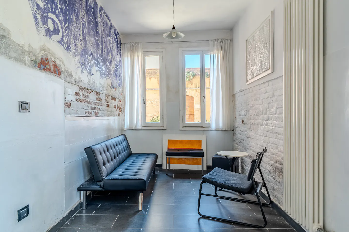 A small room with a black futon, chair, and bench. The walls are partially exposed brick and painted white. Two windows let in natural light.
