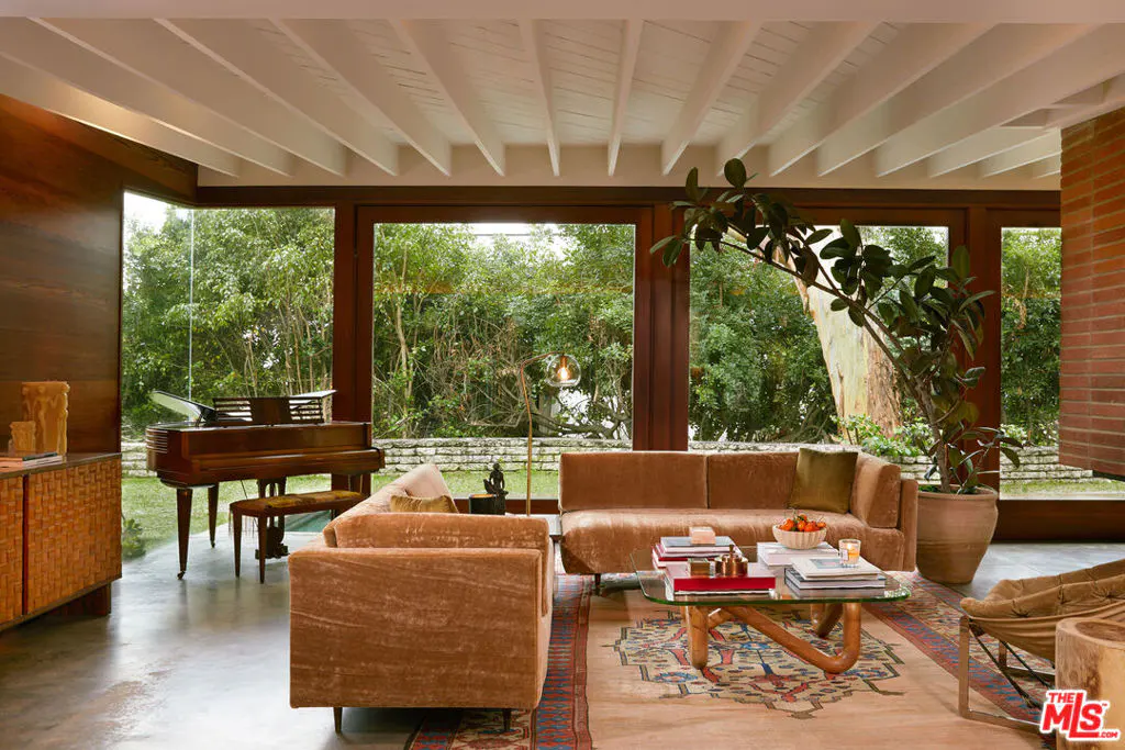 Living room with brown velvet sofas, glass table with books, and a piano by large windows overlooking greenery.