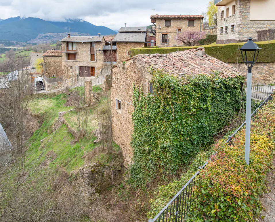 Stone houses with red tile roofs sit on a hillside. Green ivy covers one building. A black lamppost stands on the right.