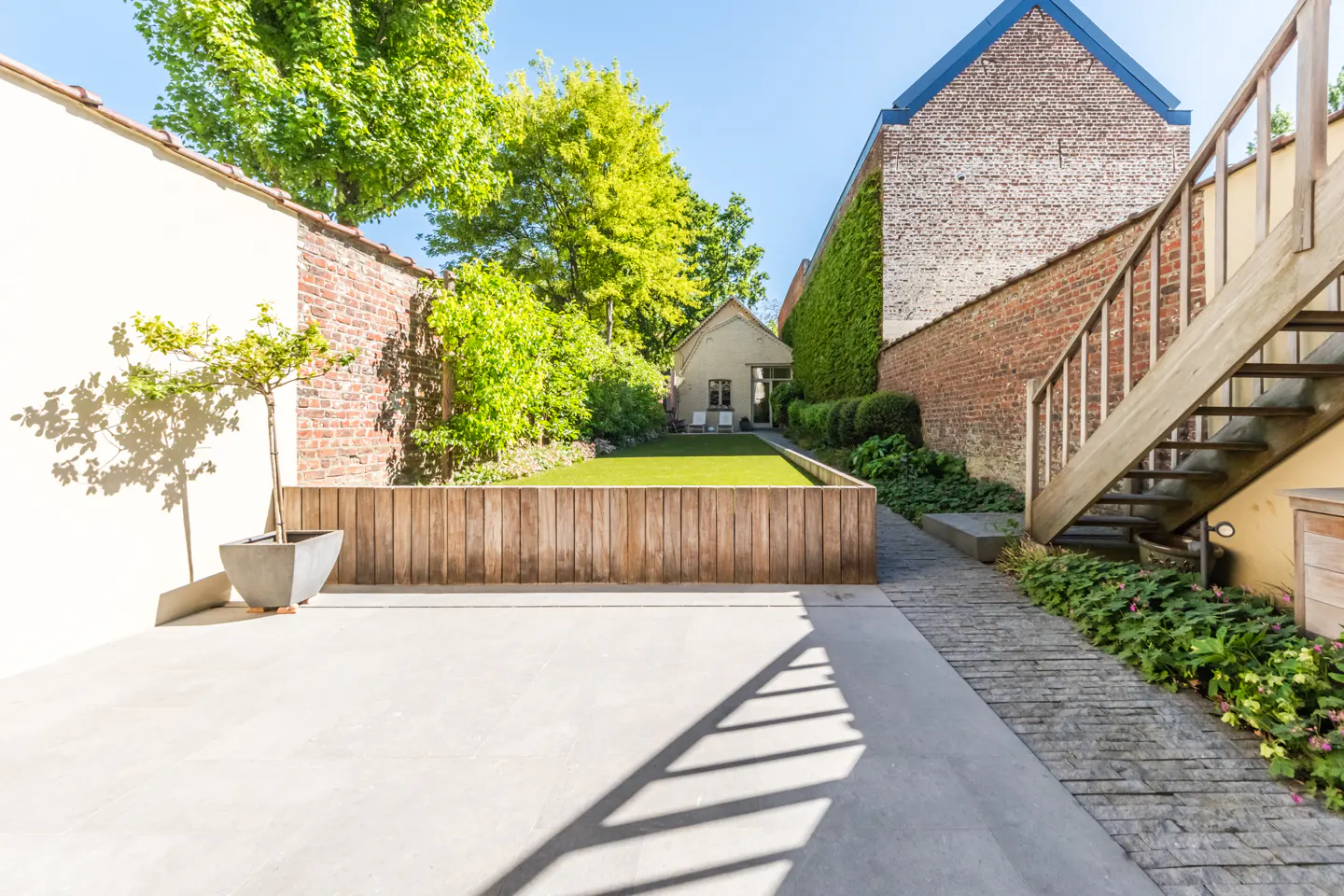 A sunny backyard with a wooden fence enclosing a green lawn, brick walls, and a wooden staircase.