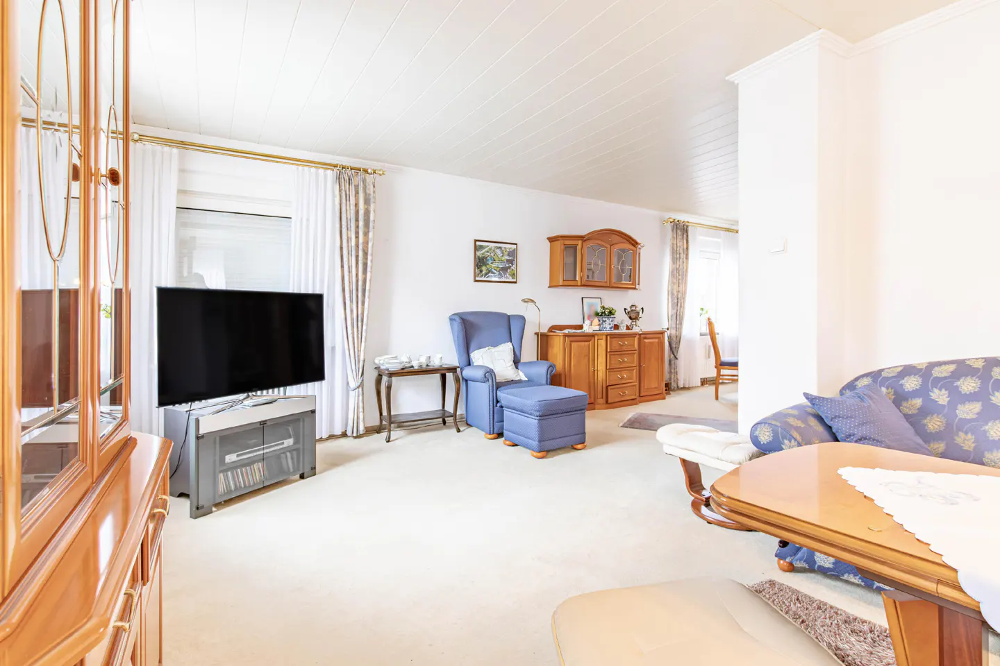 Living room with beige carpet, white walls, blue armchair, wood furniture, and a large TV. Curtains frame the windows.