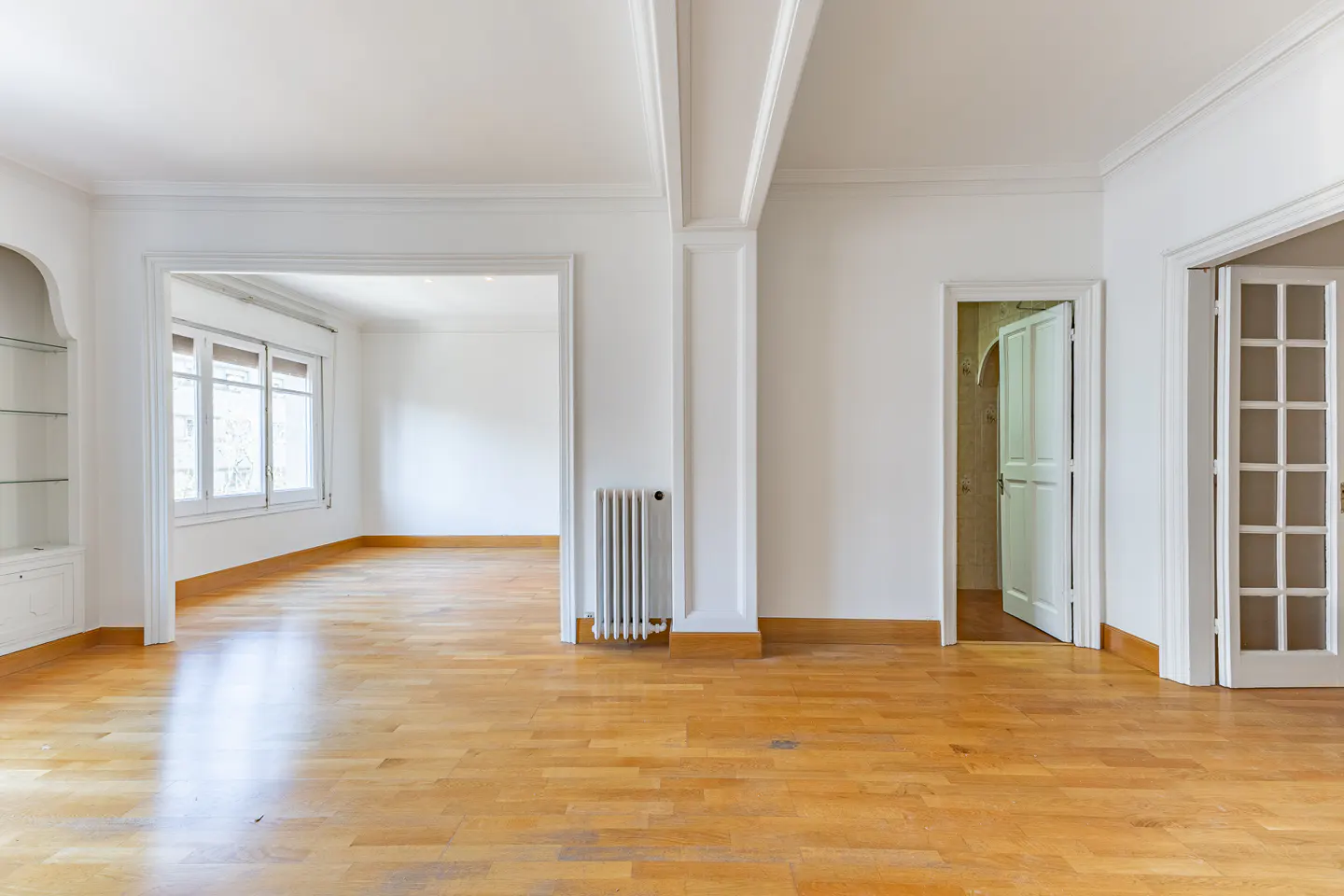 Bright, empty room with hardwood floors, white walls, and trim. Doorways lead to other rooms. A radiator is visible.