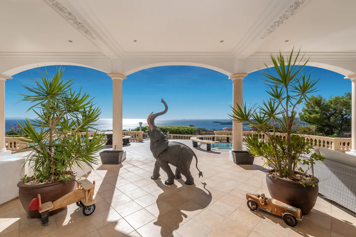 Luxury patio with ocean view, framed by white columns. A gray elephant statue stands center, flanked by potted plants and toy cars.