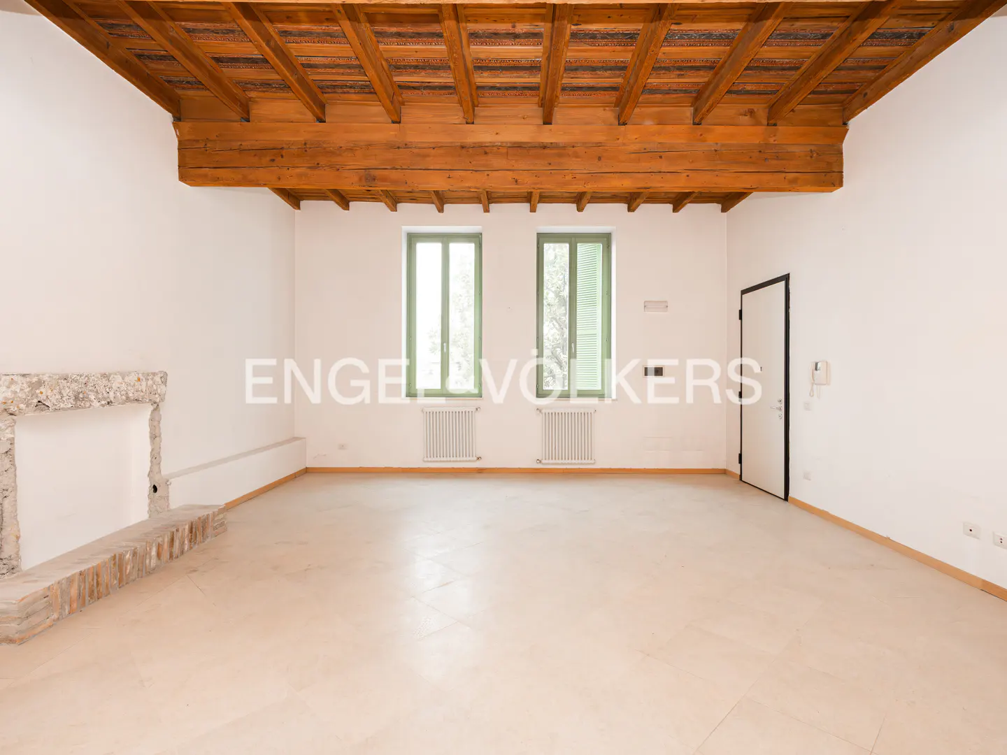Empty room with white walls, light beige floor, and exposed wood beam ceiling. Two green-framed windows and a stone fireplace are visible.