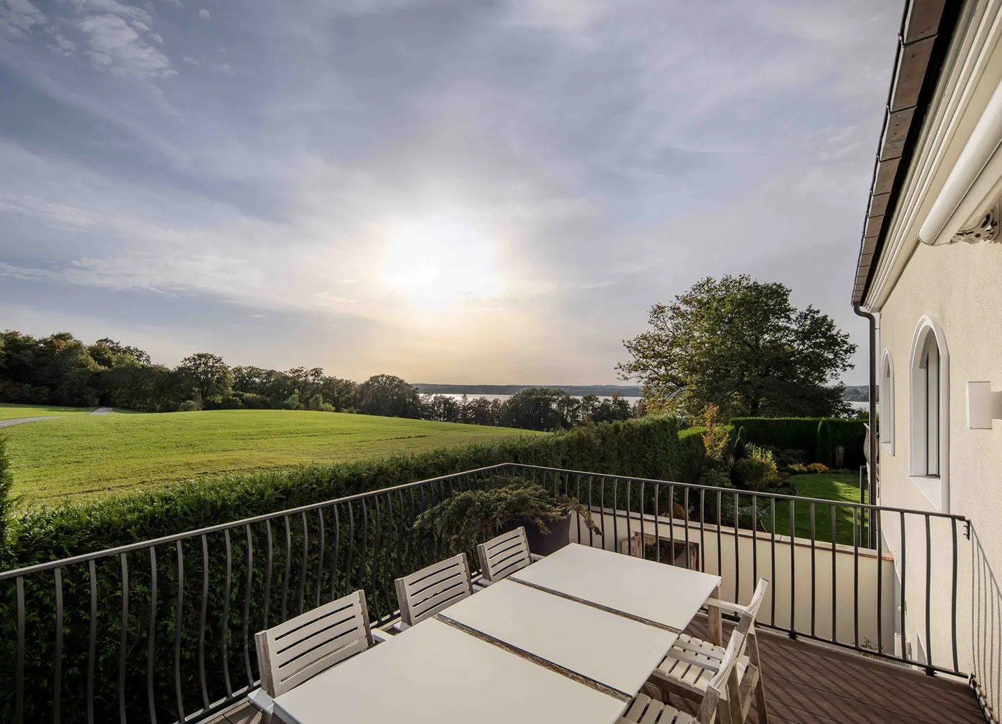 Outdoor patio with white table and chairs overlooking green lawn, trees, and lake under a cloudy sky.