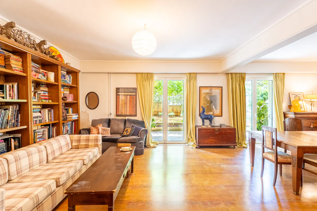 Bright living room with a large bookshelf, sofas, a wooden table, and French doors leading to a garden.