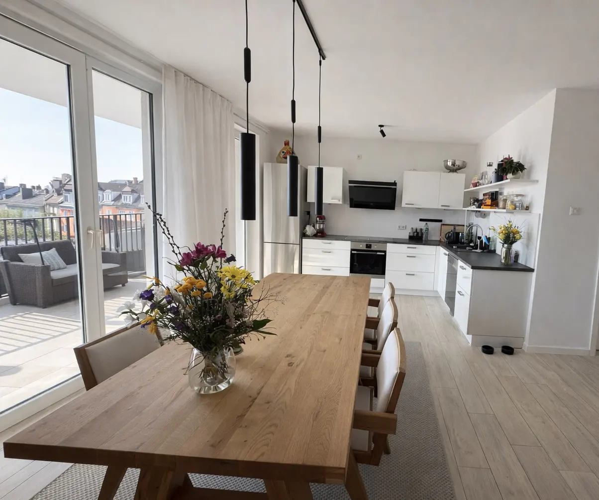 Bright, modern kitchen and dining area with a wooden table, chairs, and a vase of colorful flowers. Balcony with outdoor furniture visible.