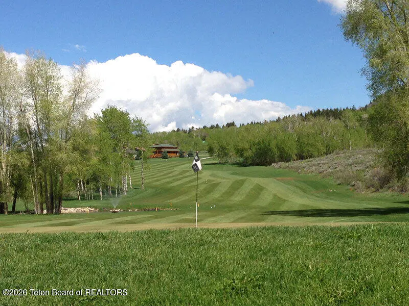 A golf course fairway with a flag in the foreground, surrounded by lush green trees and a distant house under a blue sky.