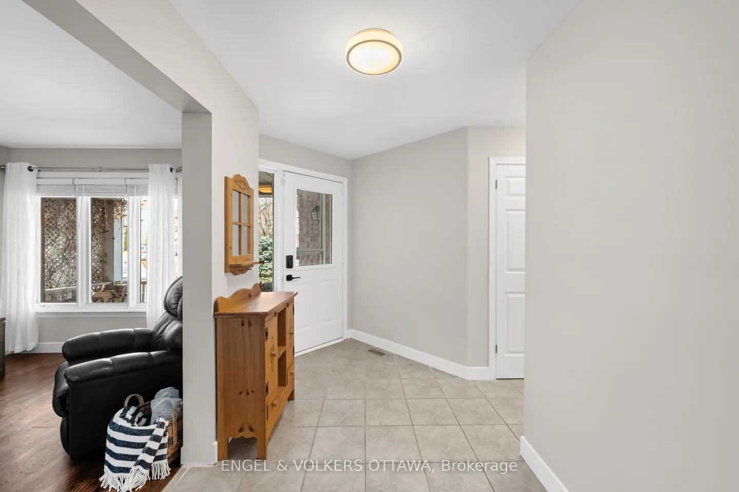 A bright foyer with a white front door, a wooden cabinet, and a glimpse into a living room with a black recliner.