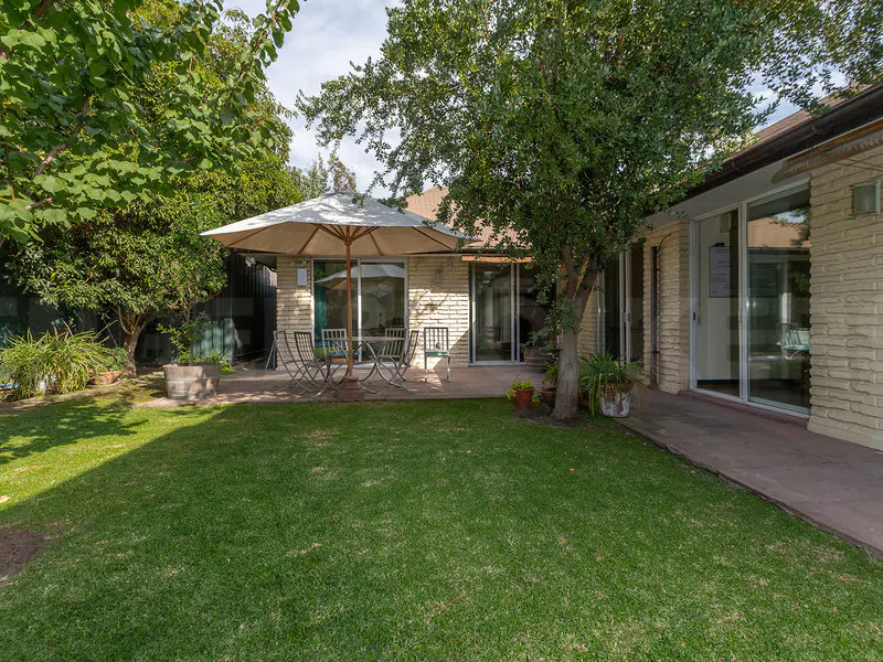 A backyard with green grass, a patio with a table and chairs under an umbrella, and a brick house.