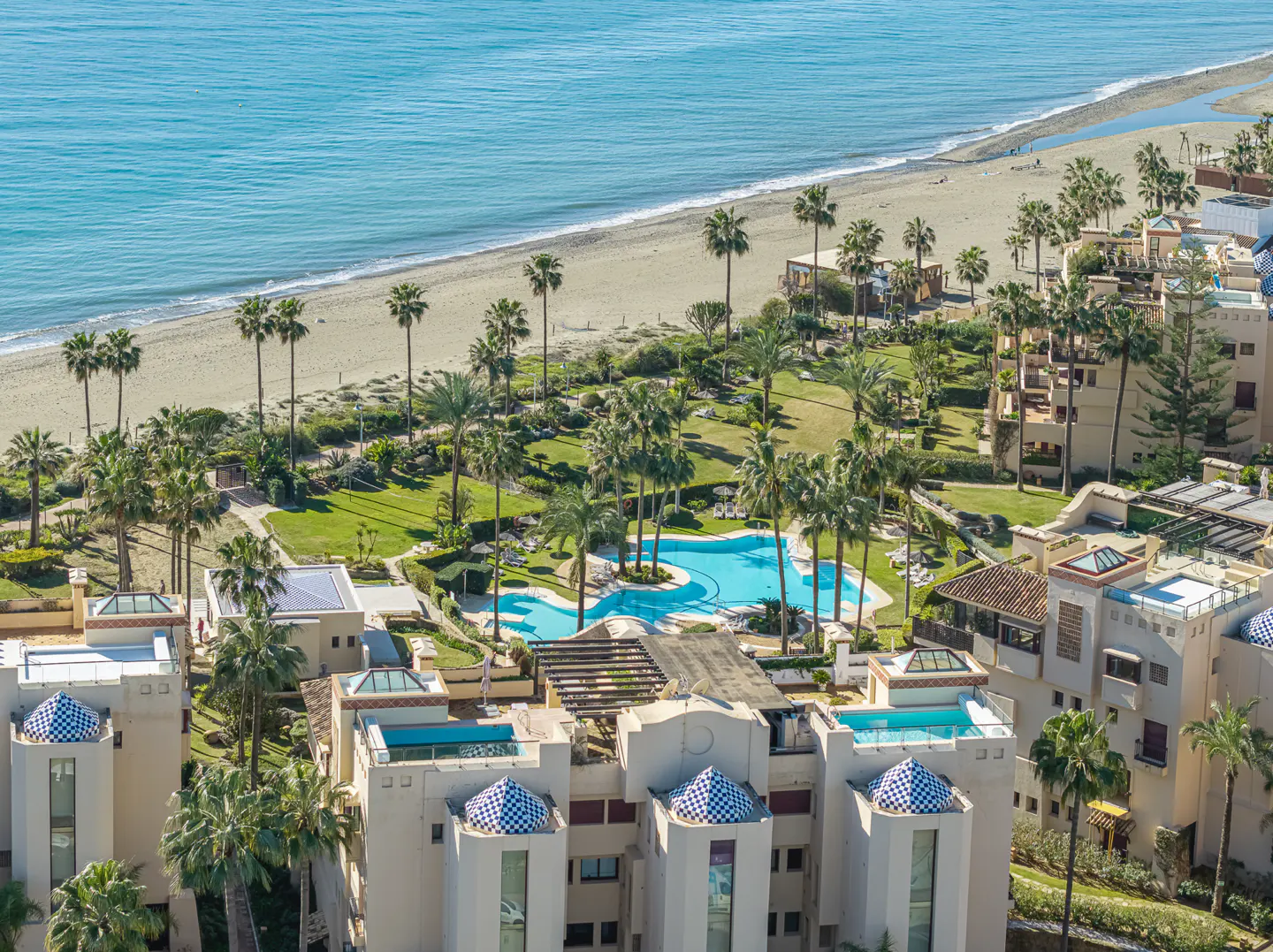 Aerial view of a beachfront resort with palm trees, pools, and white buildings with blue-tiled domes. Sandy beach and turquoise ocean in the background.