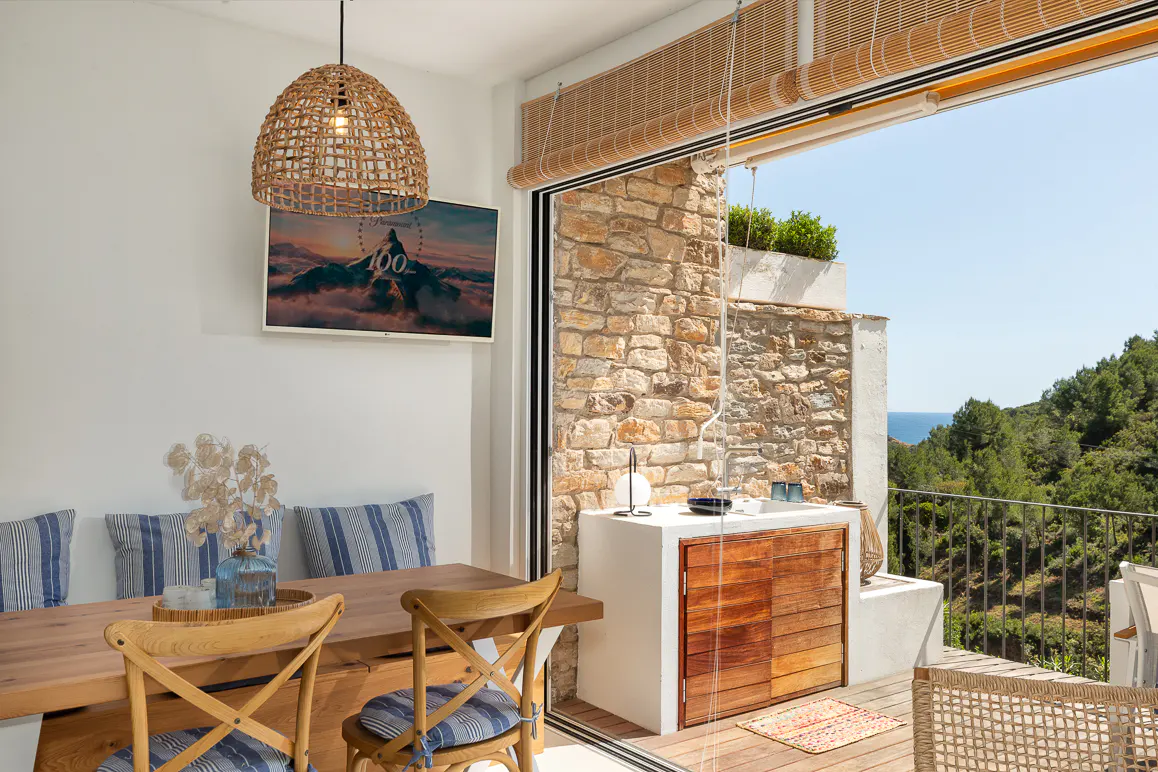 Bright dining area with a wooden table, blue striped cushions, and a woven pendant light. Sliding glass doors open to a stone patio with forest and ocean views.
