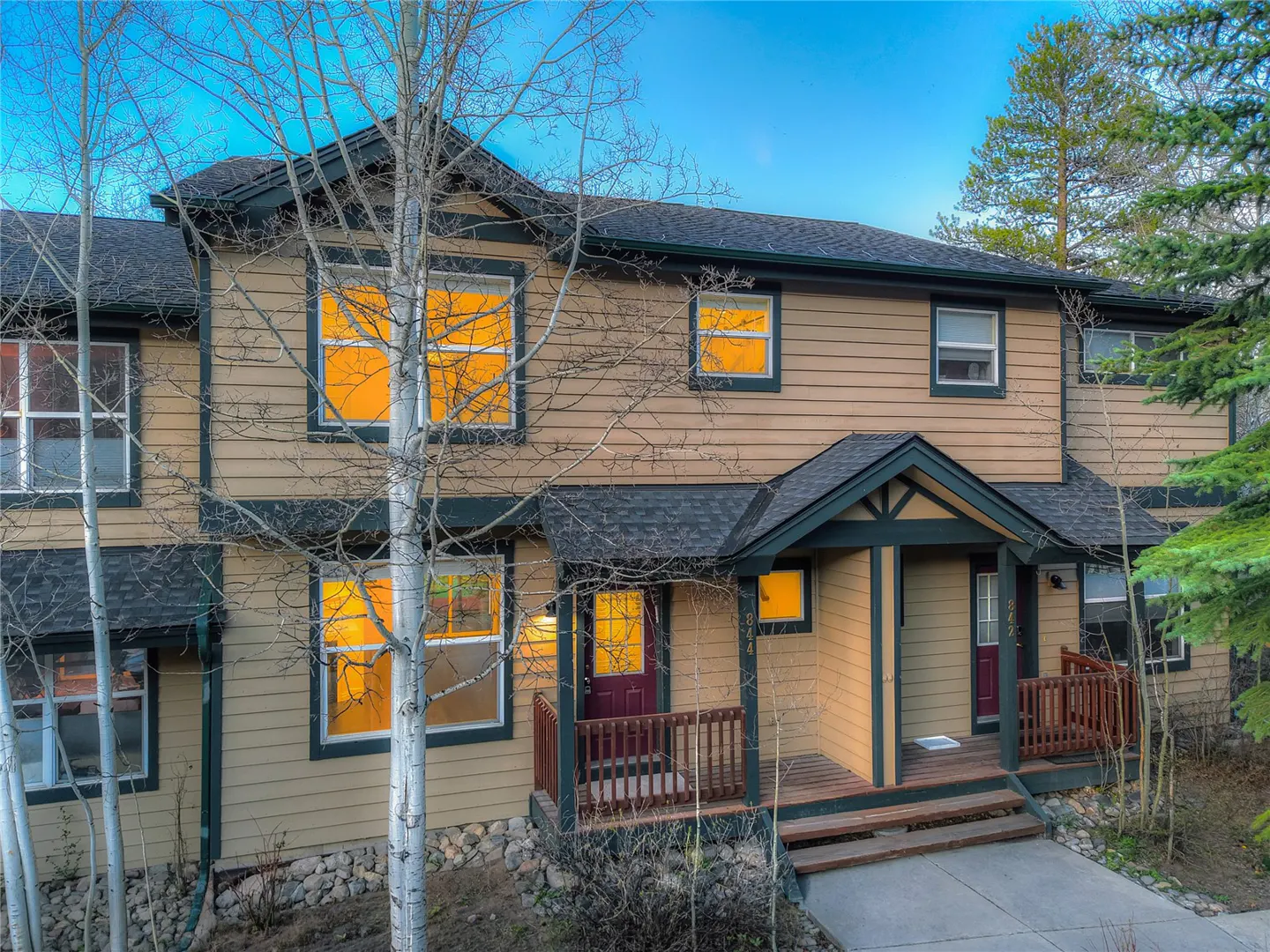 Two-story tan townhouse with dark trim and a dark roof. Windows glow with warm light. Bare trees frame the building.