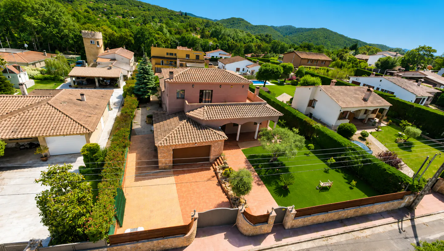 Aerial view of a salmon-colored house with a brown tile roof, green lawn, and a backdrop of lush green mountains.