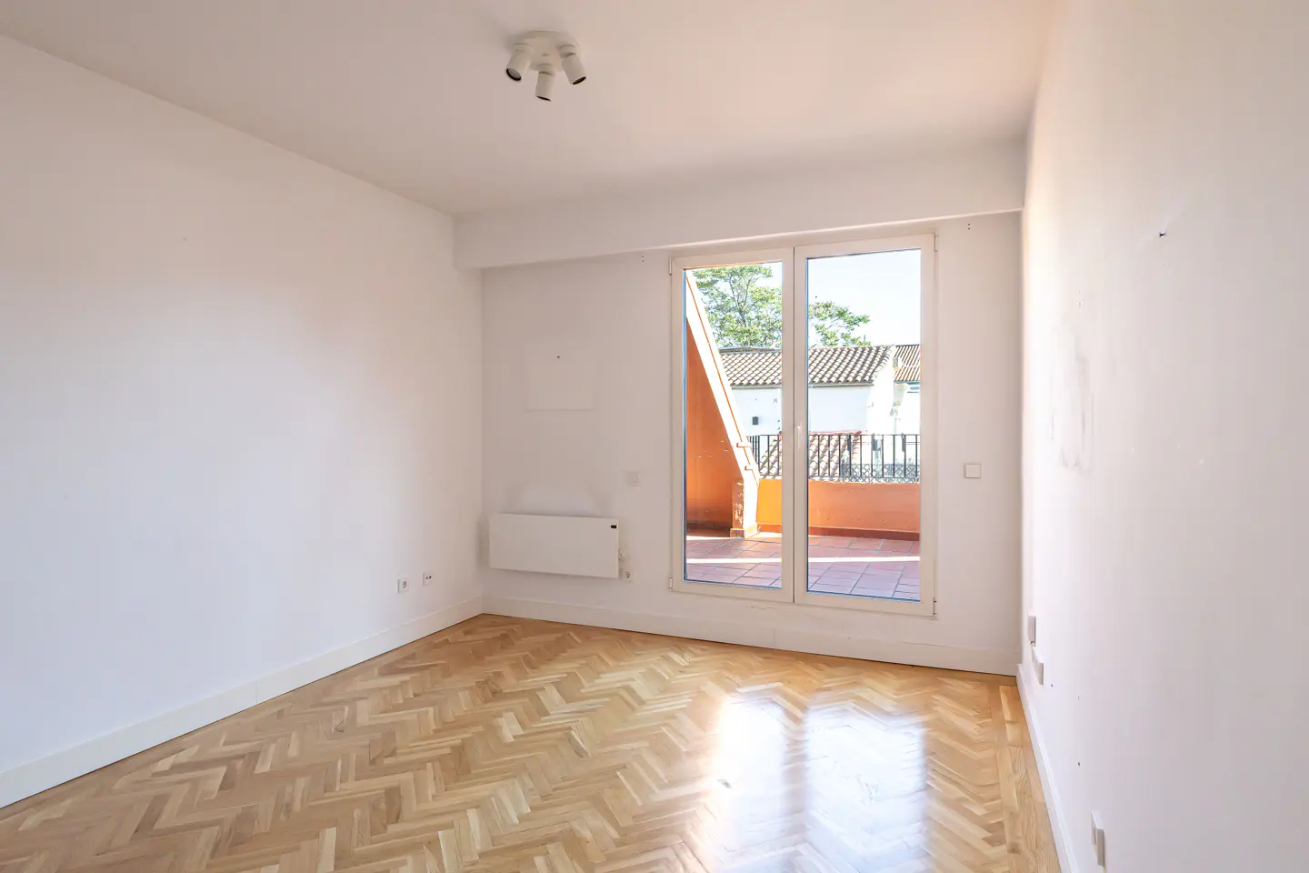 Bright, empty room with herringbone wood floor, white walls, and a sliding glass door to a balcony with terracotta tiles.