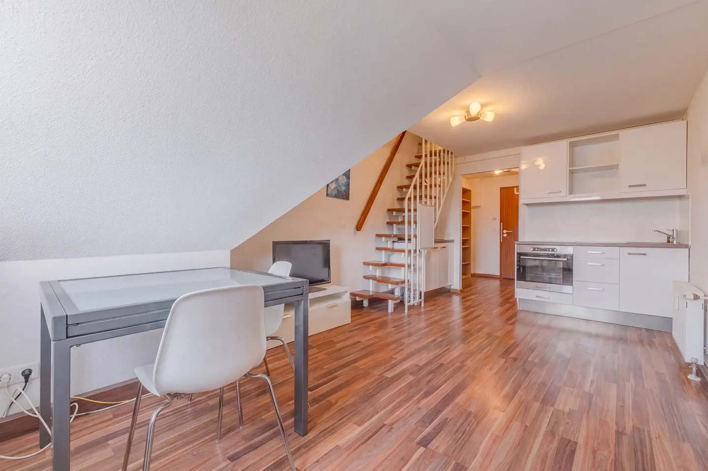 Open-concept apartment with wood floors, white walls, and a sloped ceiling. A table and chairs sit near a TV and stairs to an upper level.